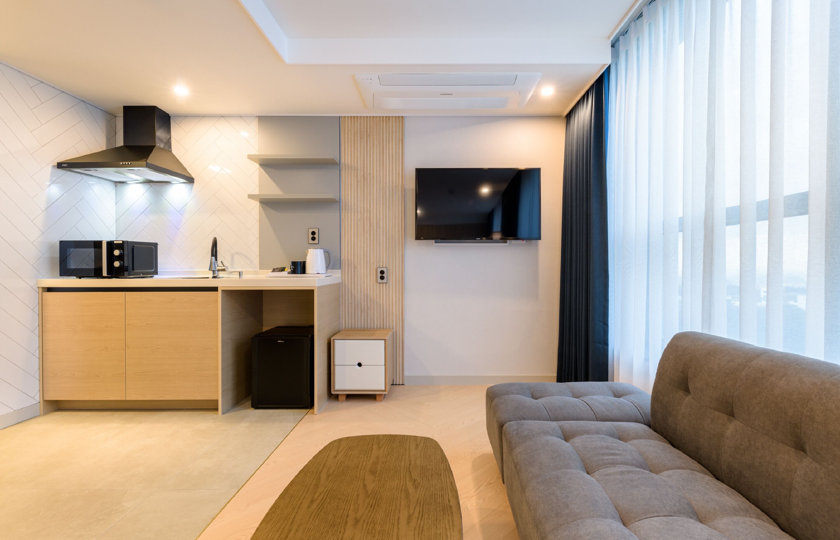 A modern living room with a sofa, a kitchen area with a microwave, a coffee maker, and a TV mounted on the wall.