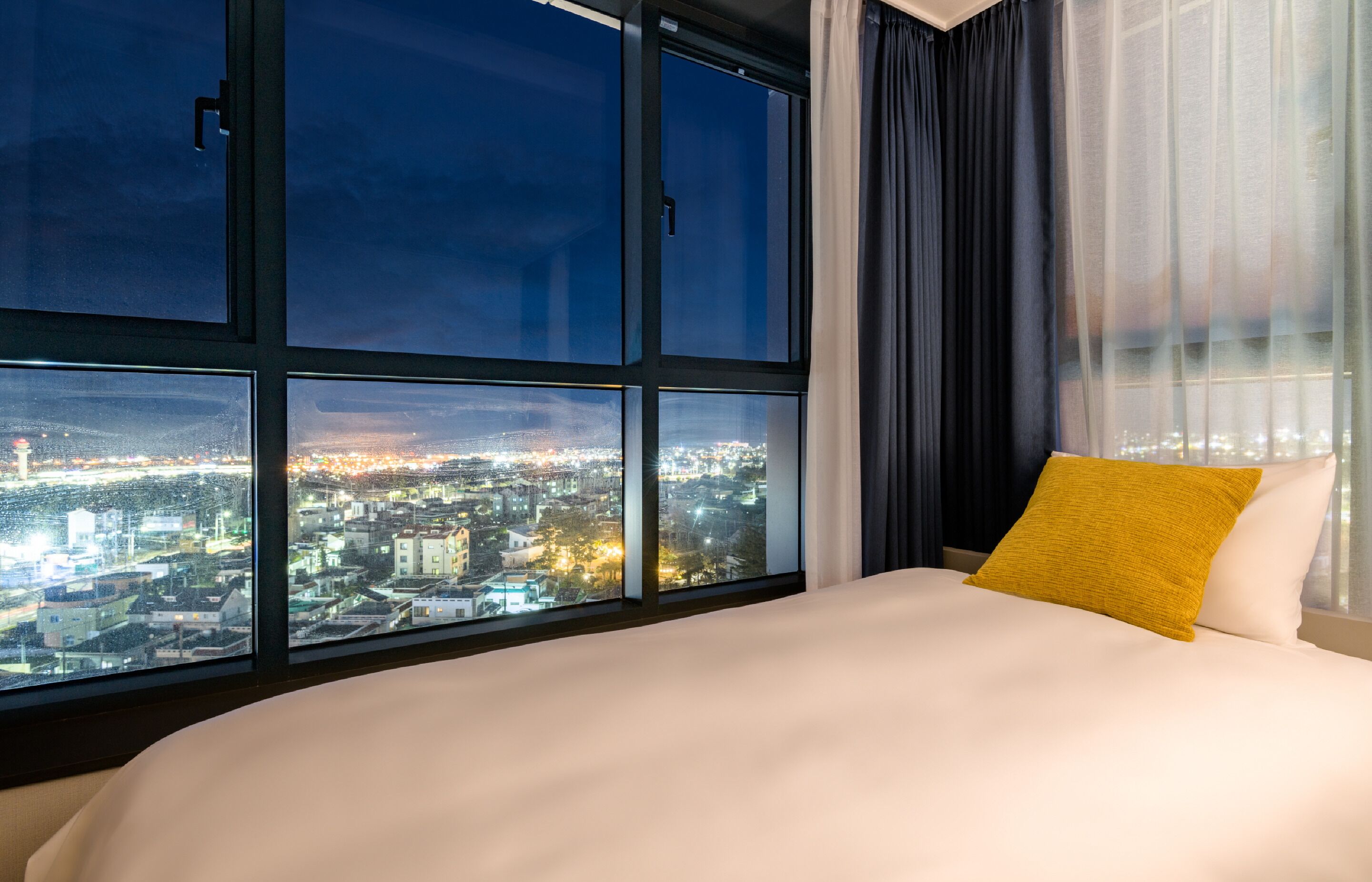 Une chambre d’hôtel dotée d’une grande fenêtre donnant sur la ville, avec une vue particulièrement agréable la nuit.