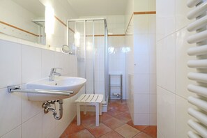 A bathroom with a white sink, a glass shower enclosure, a small white stool, and a radiator.