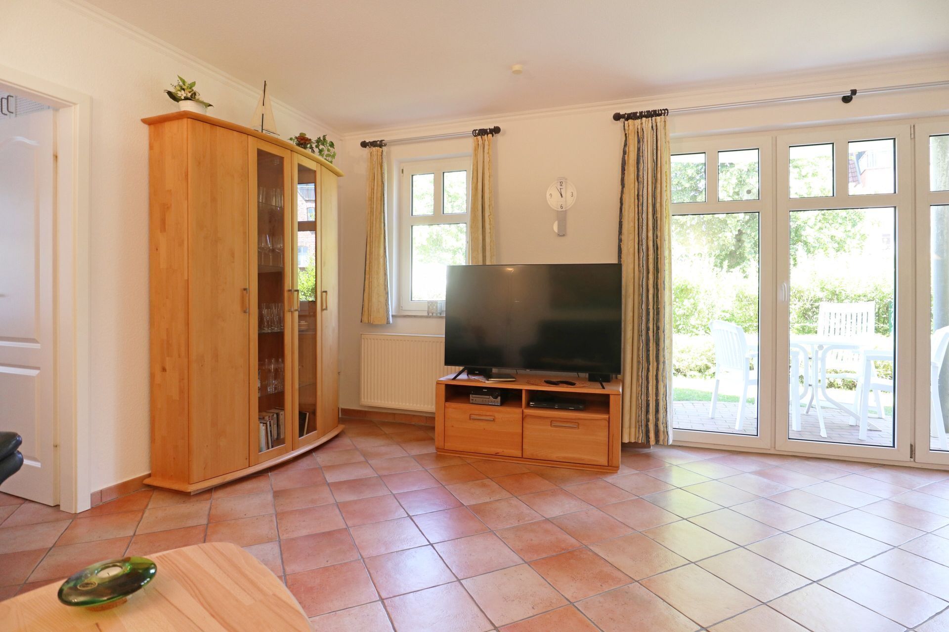 A living room with a wooden cabinet, a television on a wooden stand, and large windows with curtains.