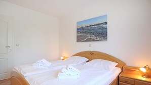 A double bed with white linens and towels, a wooden headboard, bedside tables with lamps, and a coastal landscape painting on the wall.
