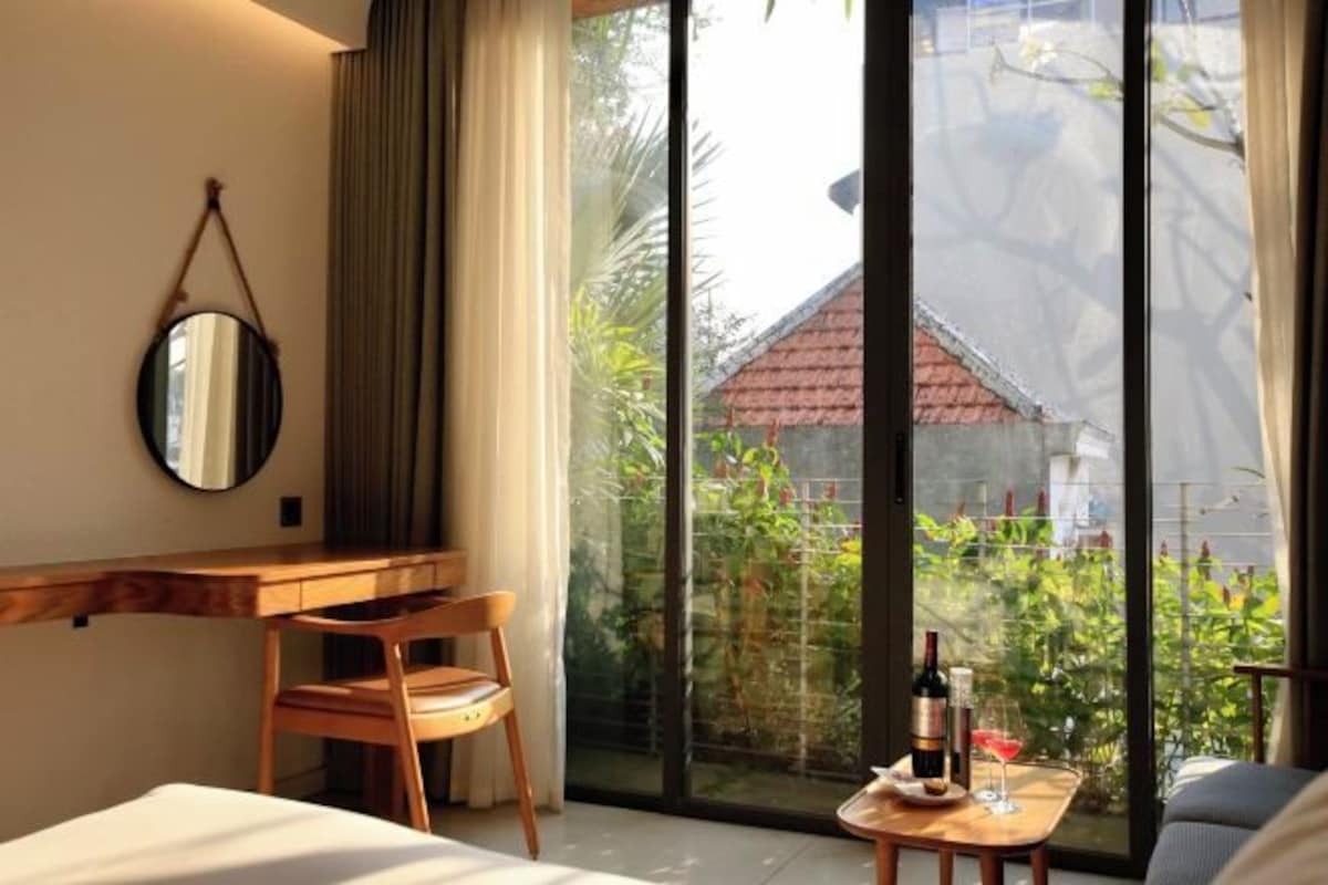 Habitación de hotel con escritorio de madera, una silla, una cama, una mesita con una botella y un vaso, y un amplio ventanal corredero que da a un jardín con un edificio de techo rojo al fondo.