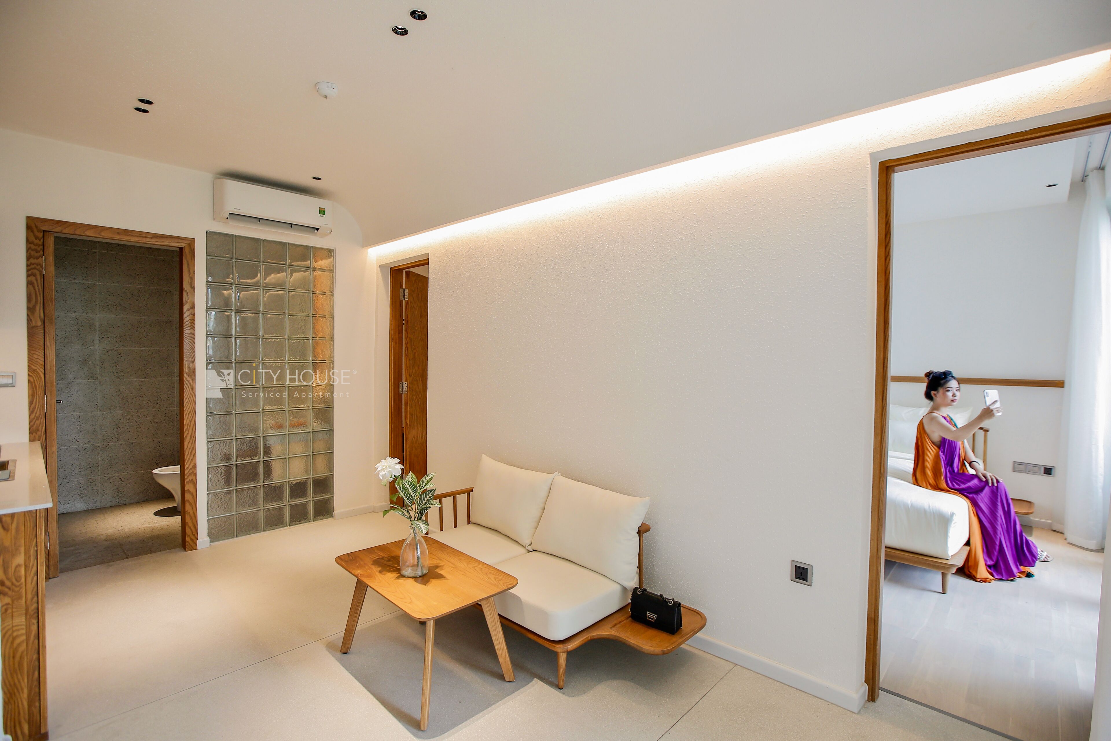 A modern bedroom with a sofa, a small table with a vase, and a mirror reflecting a person.