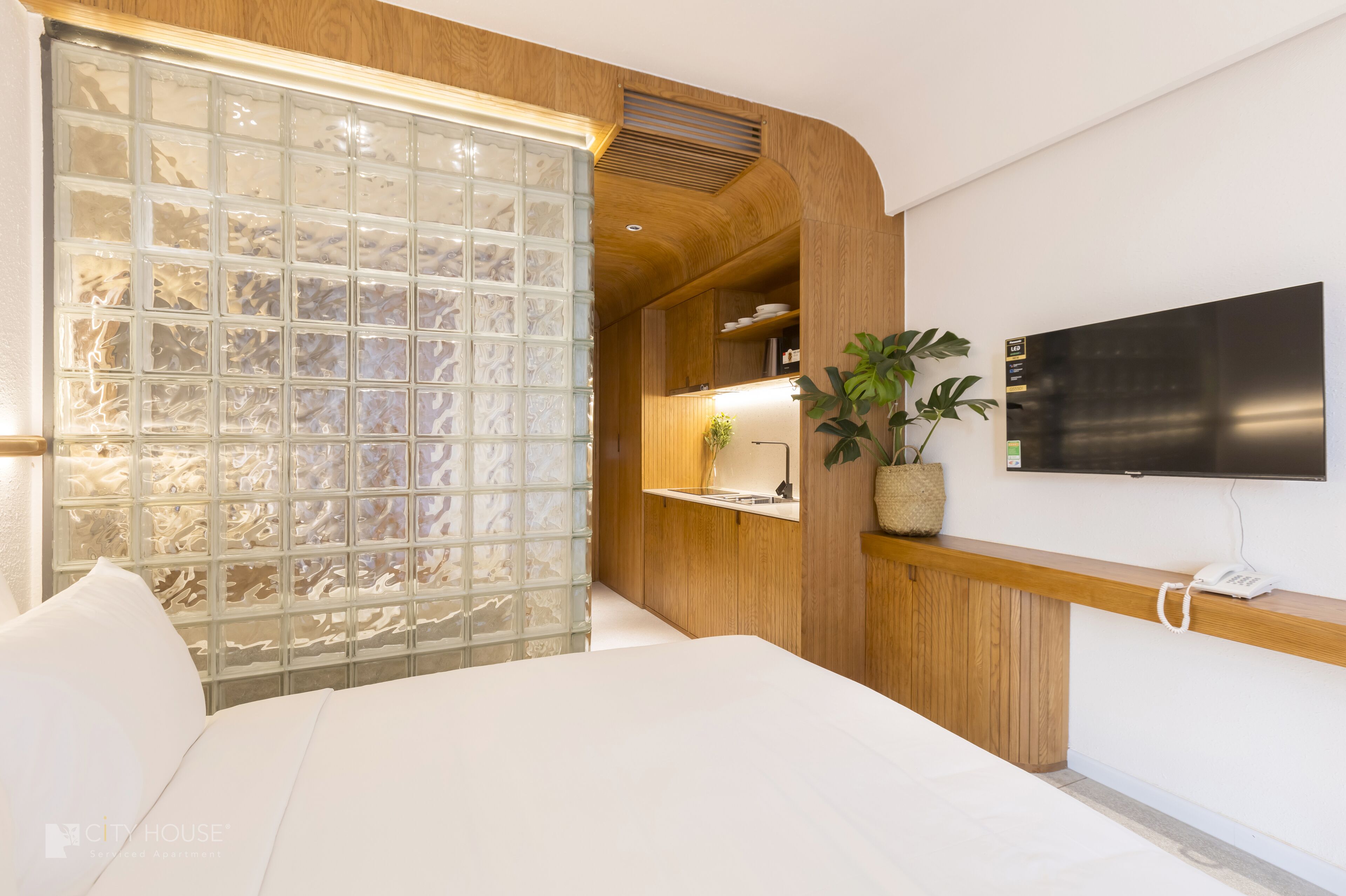 Una habitación de hotel moderna con una cama grande, un separador de vidrio, un banco de madera, un televisor montado y una planta en maceta.