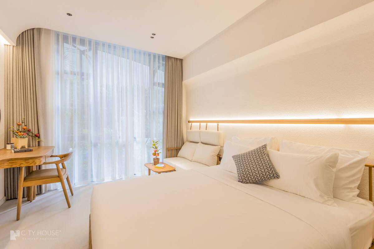 A modern hotel room with a large bed, a desk, and a chair. There is a window with curtains, a small table with a plant, and a headboard with built-in lighting.