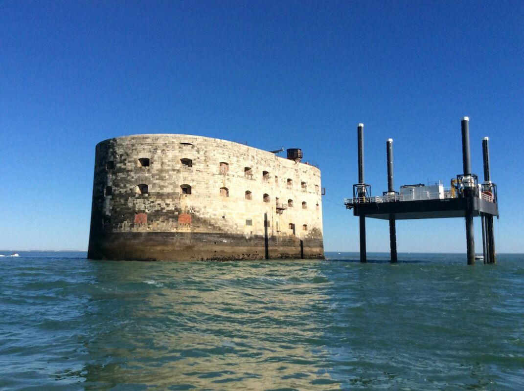 A cylindrical stone structure with a flat top, partially submerged in water, supported by a pier with multiple vertical posts.