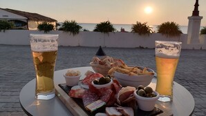 Une table avec deux verres de bière, une planche de charcuterie avec différentes sortes de viandes et d’olives, et une bouteille de spiritueux, le tout installé à l’extérieur, avec un coucher de soleil en arrière-plan.