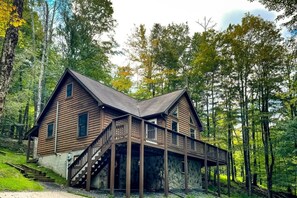 A two-story wooden cabin with a stone foundation, surrounded by a forest.