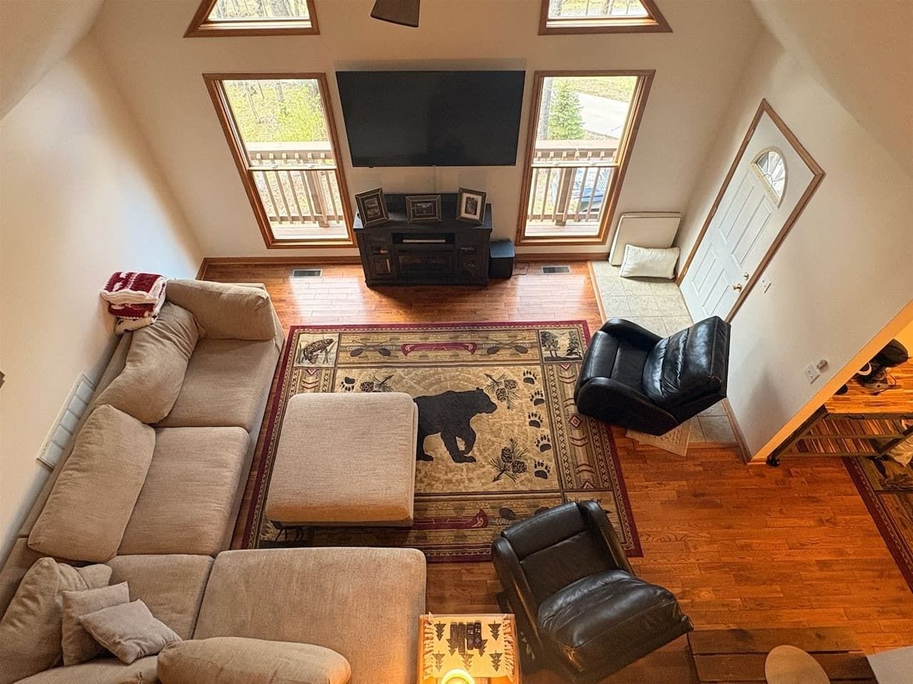 A living room with a sofa, ottoman, and a black leather chair. A flat-screen TV is mounted on the wall, and there is a bear rug on the floor.