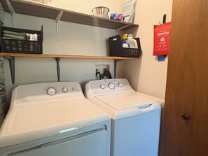 A laundry area with two stacked washers and dryers, a shelf above with cleaning supplies, and a fire blanket hanging on the wall.