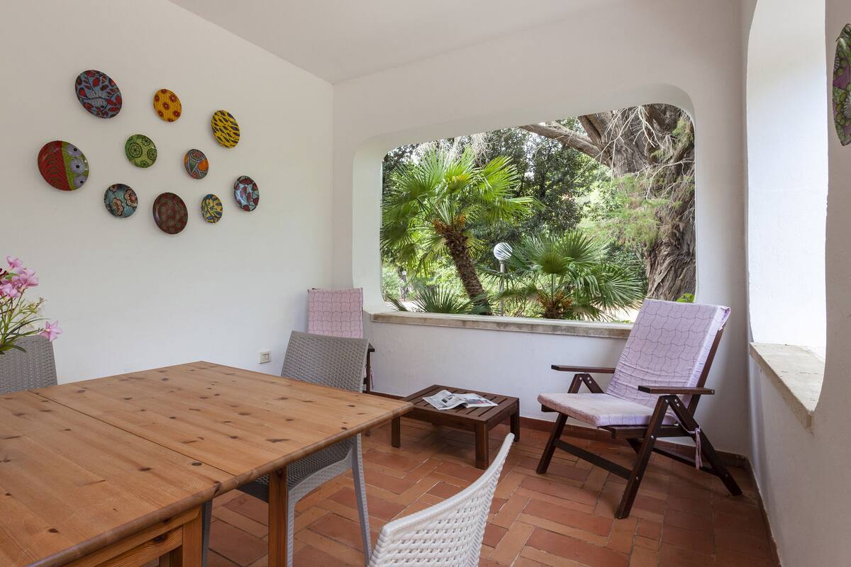 A dining area with a wooden table, chairs, a vase of flowers, and a window with a view of greenery.
