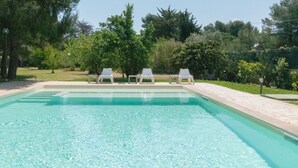 Piscina retangular com água azul cristalina, rodeada por um deck revestido com azulejos. Três cadeiras de espreguiçadeira brancas estão alinhadas na borda da piscina.