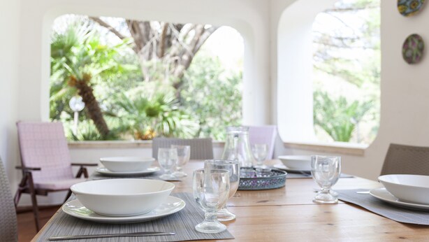 Mesa de jantar posta com pratos brancos, tigelas e copos de vinho transparentes, com vista para vegetação exuberante através de grandes janelas.