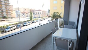 Ein Balkon mit Tisch und Stühlen, der auf eine Straße mit parkenden Autos und Gebäuden im Hintergrund blickt.