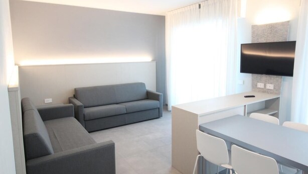 Ein modernes Hotelzimmer mit grauem Sofa, einem kleinen Esstisch und einem wandmontierten Flachbildfernseher.