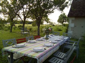 Un espacio exterior con comedor, mesa dispuesta para seis personas, sillas alrededor, zona verde con árboles y un edificio al fondo.