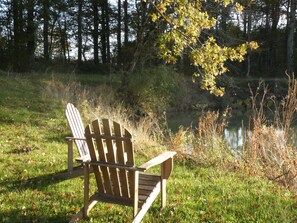 A wooden Adirondack chair in a grassy area with a pond and trees in the background.