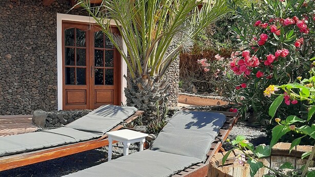 A patio with wooden lounge chairs and a small table, surrounded by a stone wall and various plants. - Volcanic lava stone house short walk to the beach,  sea and mountain views (Arrieta, Lanzarote)