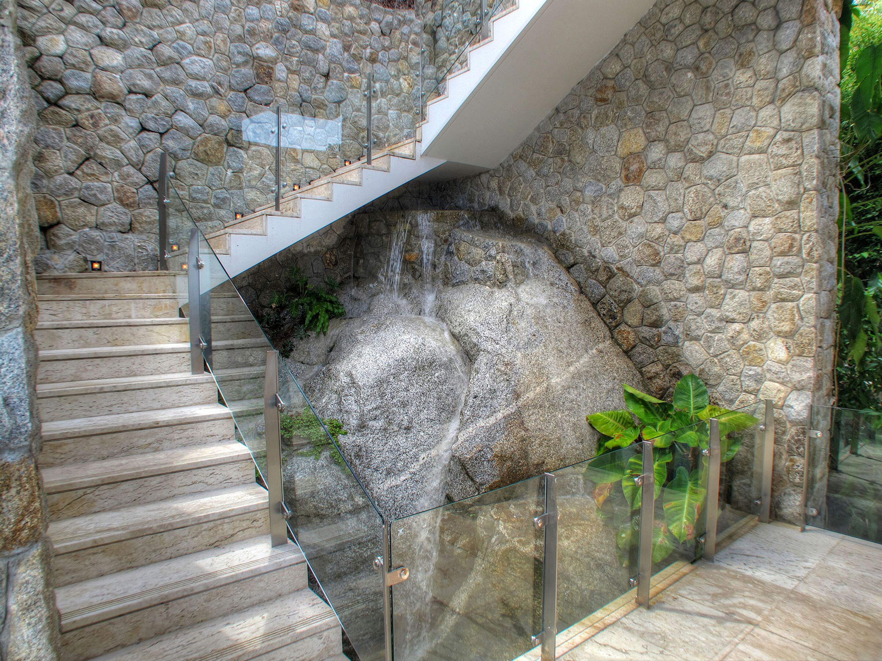A modern staircase with glass railings leading to a stone wall with a water feature.