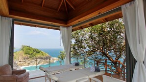 Una habitación iluminada por el sol con techo de madera, una camilla de masaje, un sillón acolchado y vistas al océano y la vegetación.