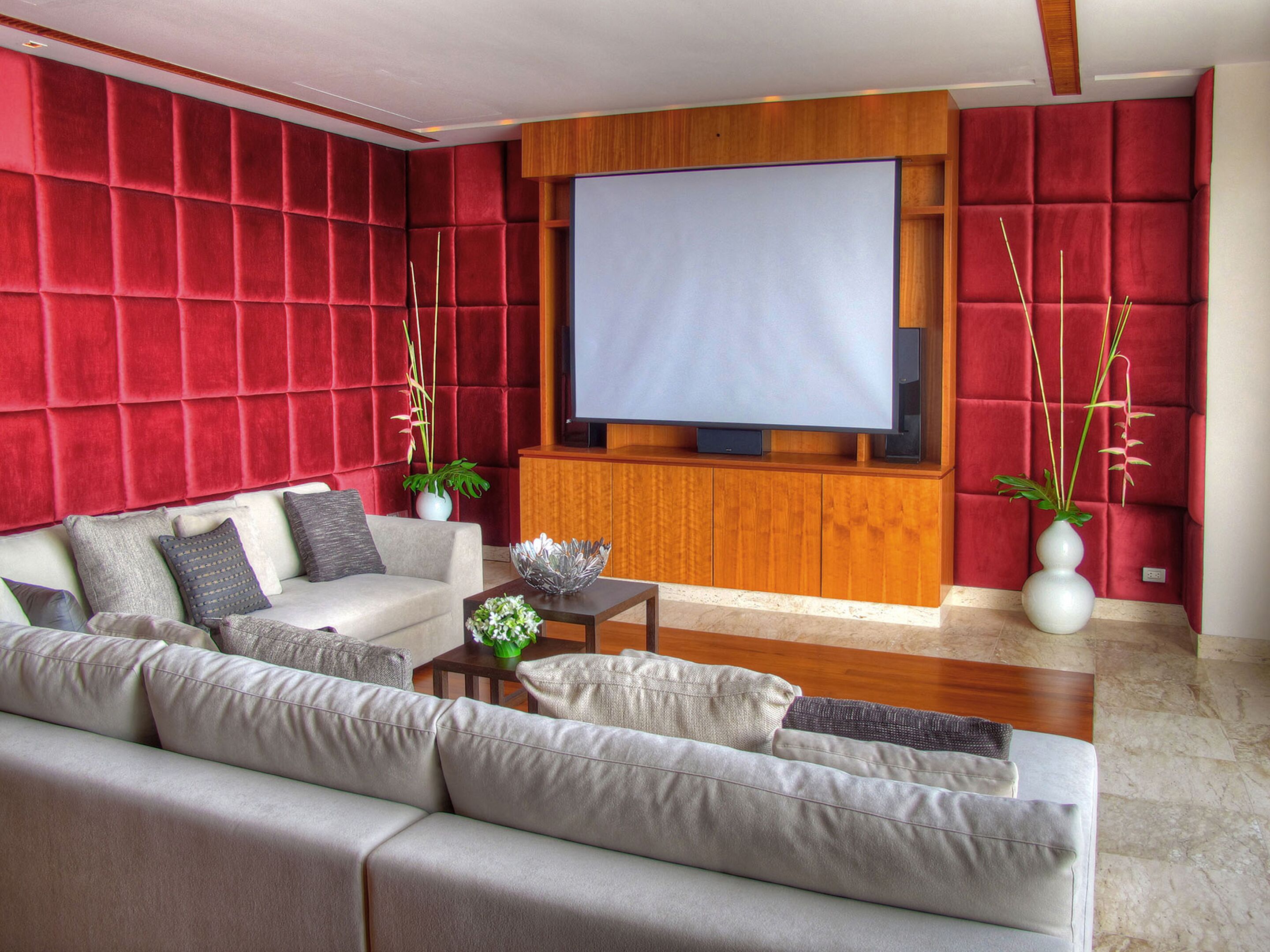 A modern living room with a large flat-screen TV, plush red upholstered walls, a grey sofa, and a wooden coffee table.