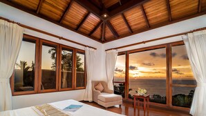 A bedroom with a large bed, a chair, a small table, and a view of the ocean at sunset. - Baan Bon Khao (Choeng Thale)