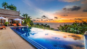 A infinity pool with a view of the ocean at sunset, lounge chairs, and a building with a tiled roof. - Baan Bon Khao (Choeng Thale)
