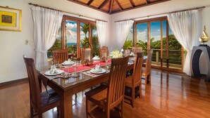 A dining area with a wooden table set for four, surrounded by chairs, with a view of the outdoors through large windows. - Baan Bon Khao (Choeng Thale)