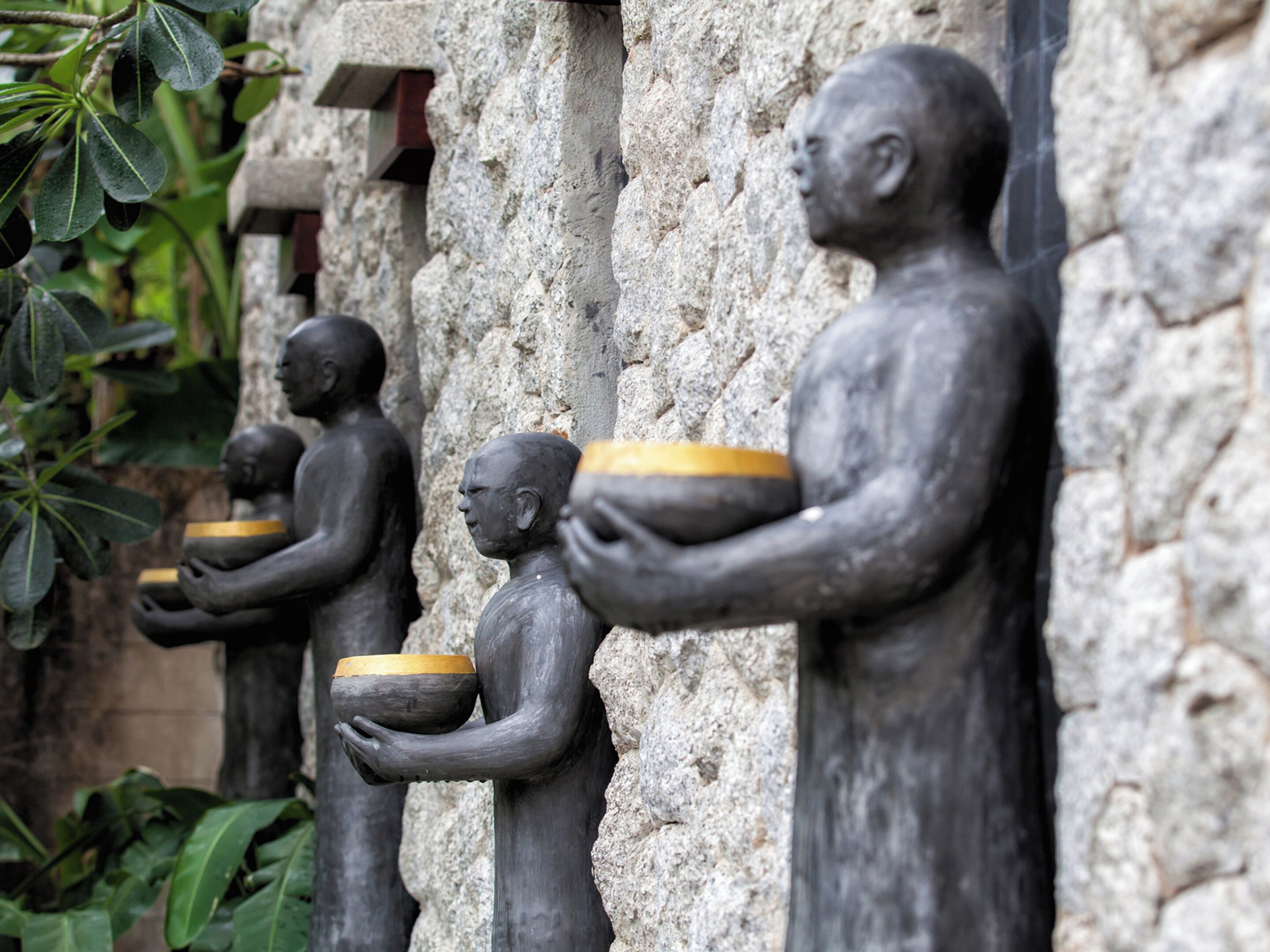 A row of stone statues holding bowls, set against a textured stone wall.