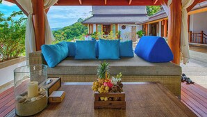 A covered outdoor seating area with a view of a traditional building and lush greenery. - Baan Surin Sawan (Choeng Thale)