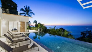 Una piscina infinita moderna con vistas despejadas al océano al atardecer, sillas de descanso y un balcón con vista al mar.