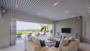 Una sala moderna con un sofá grande, una mesa de centro y un televisor montado en la pared. Hay una puerta corrediza de vidrio que da a un patio exterior con sillas y una mesa.