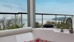 A modern bathroom with a bathtub, a large window offering a view of the sea and hills, and a decorative vase on the windowsill. - Villa Napalai (Choeng Thale)