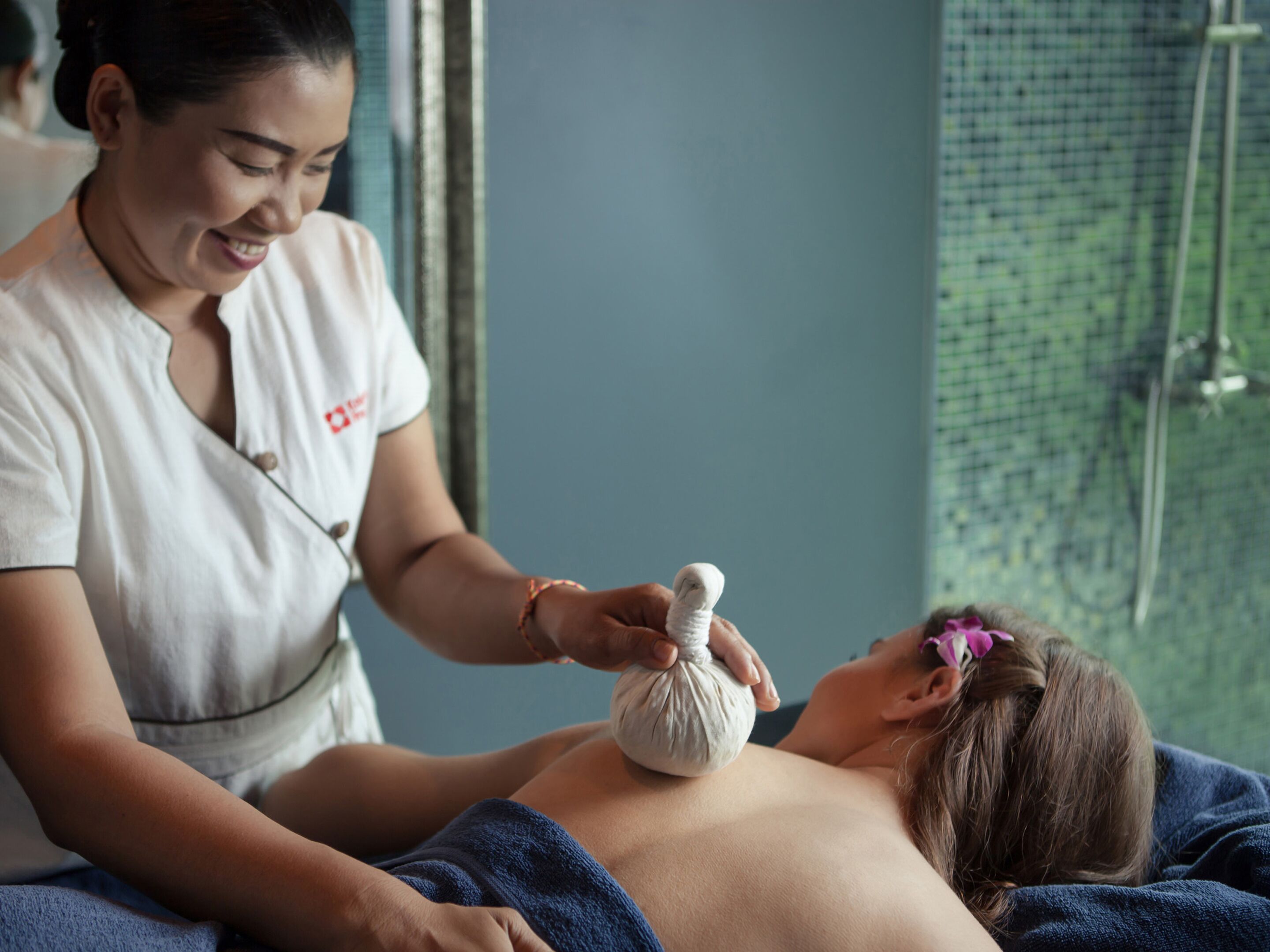 Een persoon ontvangt een massage, het lichaam bedekt met een handdoek, terwijl een massagetherapeut een behandeling uitvoert, en er is een raam met uitzicht op groen.