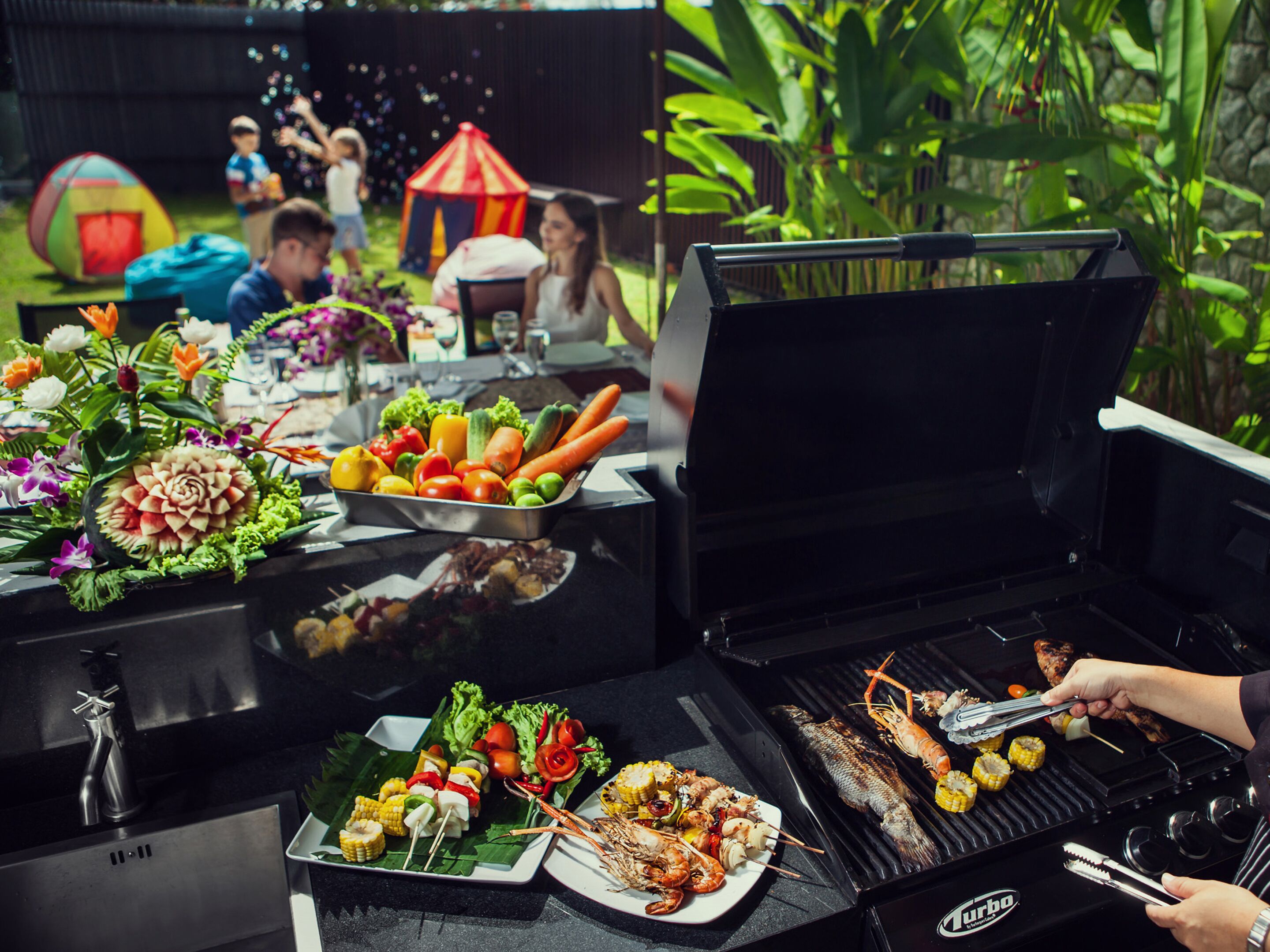 A barbecue setup with various grilled foods and a buffet table with vegetables and fruits.