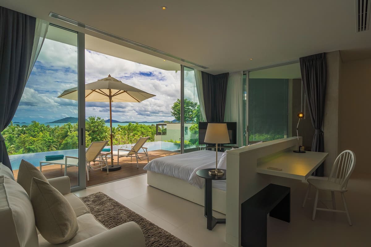 A modern hotel room with a large bed, a desk, a chair, a TV, and a view of a lush landscape and ocean.