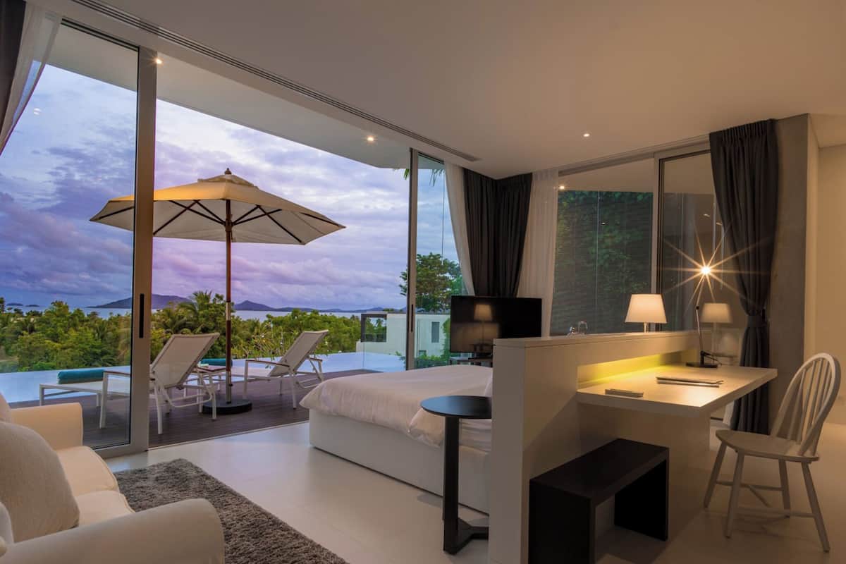 A modern hotel room with a large window, a bed, a desk, and a lounge area with a view of the sea.