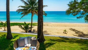A wicker lounge chair with cushions and a hat on the grass near a beach with palm trees and a clear view of the ocean.