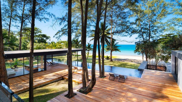 A modern outdoor deck with wooden flooring, surrounded by tall trees and overlooking a beach.