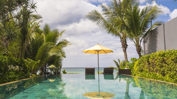 Ein Infinity-Pool mit klaren blauen Fliesen, umgeben von üppiger Vegetation und Palmen, mit Blick auf das Meer im Hintergrund.