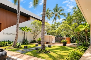 Un bâtiment moderne, sur lequel on peut lire l’inscription « NANDANA », et un jardin bien entretenu, agrémenté de palmiers et de diverses plantes.