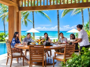 Un groupe de personnes est assis à une table en plein air, devant un panorama sur l’océan et les palmiers.