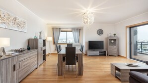 A modern living room with a dining area, wooden flooring, a large window with curtains, a flat-screen TV, and a city view. - 2-Bedroom Apartment with Rooftop Pool, Village Marina, Olhão (Olhão)