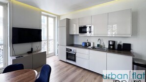 A modern kitchen with white cabinets, a built-in oven, a microwave, and a coffee maker. There is a dining area with a wooden table and blue chairs. - Urban Flat 60 - Sublime Parisian Flat in Paris (París)