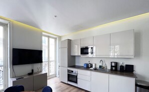 A modern kitchen with white cabinets, a built-in oven, a microwave, and a coffee maker. There is a dining area with a wooden table and blue chairs.