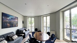 A modern living room with a large window, a sofa, a dining table, and a cityscape painting. - Urban Flat 60 - Sublime Parisian Flat in Paris (París)