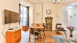 Ein Essbereich mit Holztisch, Stühlen und einer Deckenleuchte. Ein Fernseher ist an der Wand montiert, ebenso ein Schrank und ein dekorativer Spiegel.