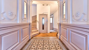 Eine prachtvolle Eingangshalle mit aufwendigen Stuckverzierungen, einem gemusterten Teppich und einer Treppe, die zu einem Aufzug führt.