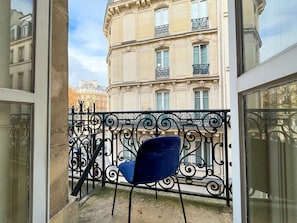 Un balcón con una silla azul, barandilla de hierro forjado negro ornamentado y vista de un edificio clásico con múltiples ventanas y balcones.