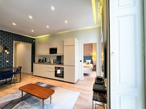 Cocina moderna con armarios blancos, microondas y cafetera. Se observa una zona de comedor con mesa y sillas. Una cama es visible al fondo.